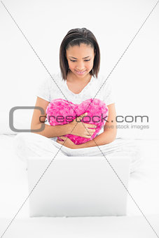 Concentrated young dark haired model cuddling a pillow and looking at a laptop