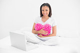 Amused young dark haired model cuddling a pillow and using a laptop