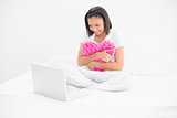 Focused young dark haired model holding a pillow and looking at a laptop