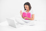 Delighted young dark haired model holding a pillow and chatting on laptop