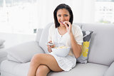 Cute young dark haired woman in white clothes eating popcorn while watching tv