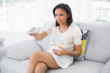 Serious young dark haired woman in white clothes changing tv chanel while eating popcorn