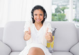 Happy young dark haired woman in white clothes listening to music and giving thumbs up