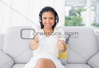 Happy young dark haired woman in white clothes listening to music and giving thumbs up
