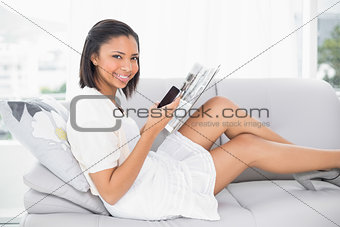 Amused young dark haired woman in white clothes reading a magazine