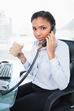 Serious young dark haired businesswoman holding a cup of coffee while answering the telephone