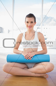 Happy fit brown haired model in sportswear sitting and holding an exercising mat