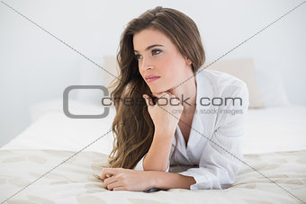 Thinking casual brown haired woman in white pajamas lying on her bed