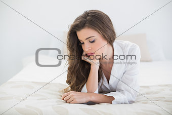 Thoughtful casual brown haired woman in white pajamas lying on her bed