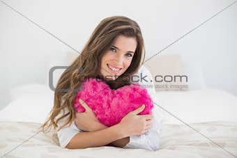Pleased casual brown haired woman in white pajamas hugging a heart shaped pillow