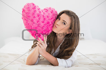 Beautiful casual brown haired woman in white pajamas kissing a heart shaped pillow