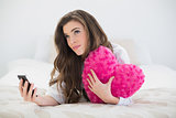 Pensive casual brown haired woman in white pajamas holding a mobile phone and a heart-shaped pillow