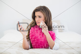 Sad casual brown haired woman in white pajamas looking at her mobile phone and crying