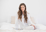 Cheerful casual brown haired woman in white pajamas sitting on her bed