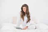 Concentrated casual brown haired woman in white pajamas using a tablet pc