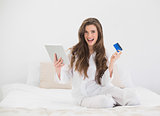 Joyful casual brown haired woman in white pajamas shopping online with her tablet pc