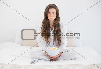 Content casual brown haired woman in white pajamas watching tv while eating popcorn