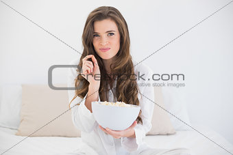 Amused casual brown haired woman in white pajamas eating popcorn