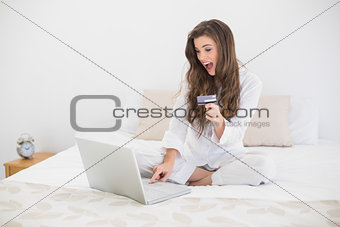 Astonished casual brown haired woman in white pajamas shopping online with her laptop