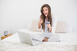 Amazed casual brown haired woman in white pajamas shopping online with her laptop