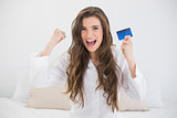 Victorious casual brown haired woman in white pajamas holding a credit card