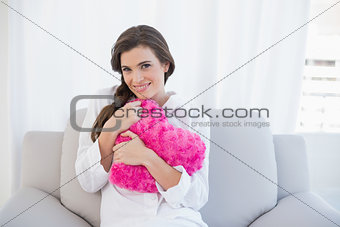 Cheerful casual brown haired woman in white pajamas hugging a heart shaped pillow