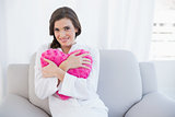Delighted casual brown haired woman in white pajamas hugging a heart shaped pillow