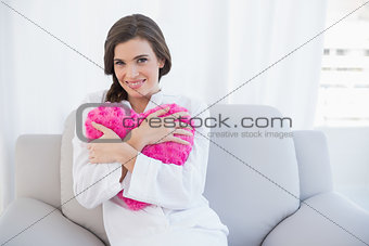 Delighted casual brown haired woman in white pajamas hugging a heart shaped pillow