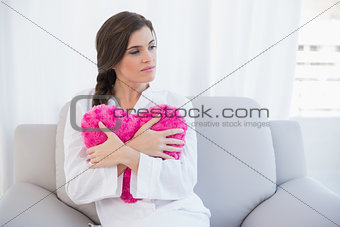 Pensive casual brown haired woman in white pajamas hugging a heart shaped pillow
