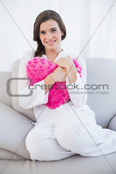 Pleased casual brown haired woman in white pajamas cuddling a heart shaped pillow