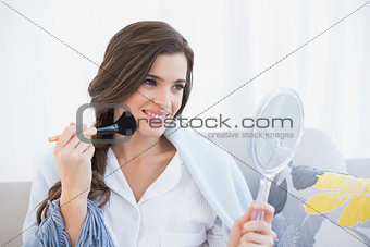 Cheerful casual brown haired woman in white pajamas applying powder on her face