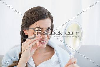 Smiling casual brown haired woman in white pajamas using an eyelash curler