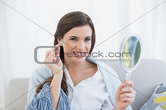 Smiling casual brown haired woman in white pajamas plucking her eyebrows