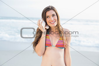 Cheerful slim brown haired model in coloured bikini listening to a shell