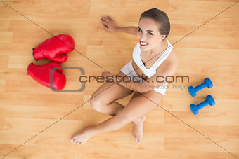 Smiling sporty brunette sitting next to red boxing gloves and dumbbells