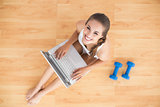 Smiling sporty brunette using a laptop and sitting next to dumbbells