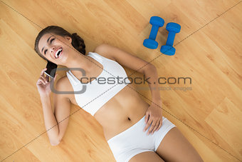 Cheerful sporty brunette using a mobile phone and lying next to dumbbells