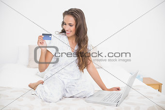 Smiling attractive brunette looking at credit card and sitting next to laptop