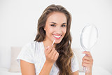 Smiling attractive brunette applying lip gloss and holding mirror