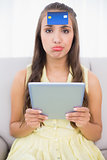 Frowning brunette with credit card on forehead holding tablet