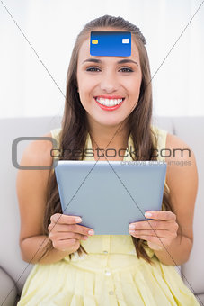 Happy brunette with credit card on forehead holding tablet