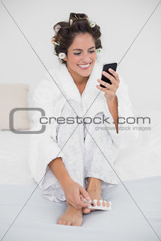 Cheerful smiling brunette applying nail polish to toenails and texting
