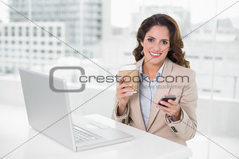 Cheerful businesswoman holding disposable cup and smartphone