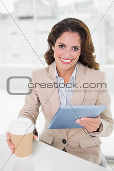 Smiling businesswoman using digital tablet at her desk holding disposable cup