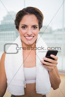 Sporty smiling brunette with towel around neck holding smartphone