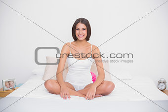 Pleased young brown haired model in white pajamas sitting on her bed