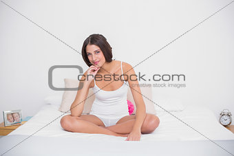 Thoughtful young brown haired model in white pajamas sitting on her bed