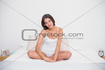 Calm young brown haired model in white pajamas sitting on her bed