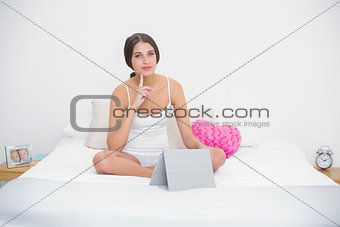Thoughtful young brown haired model in white pajamas typing on a tablet pc