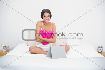 Content young brown haired model in white pajamas hugging a heart-shaped pillow